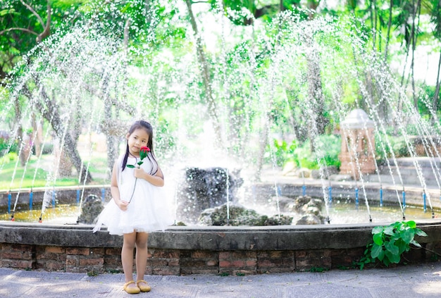 Niña asiática linda en vestido blanco sosteniendo una rosa roja en el parque