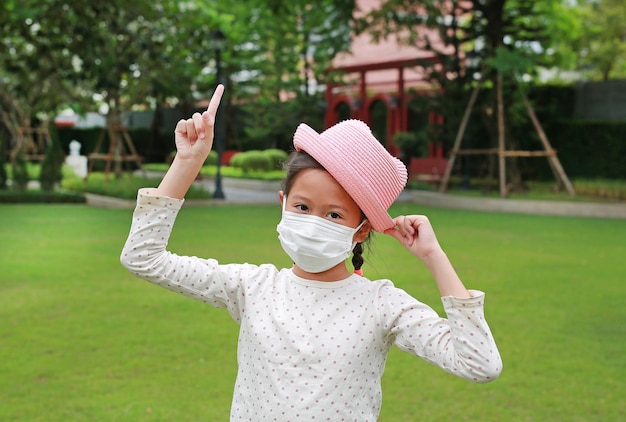 Niña asiática con máscara médica y sombrero de paja con el dedo índice apuntando hacia arriba debido a covid19 en el jardín