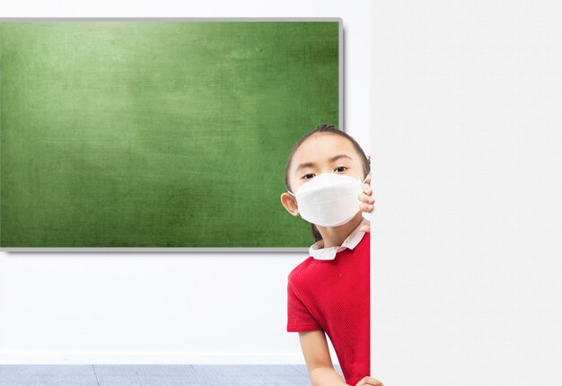 Niña asiática con la máscara de pie junto a la pared en el aula