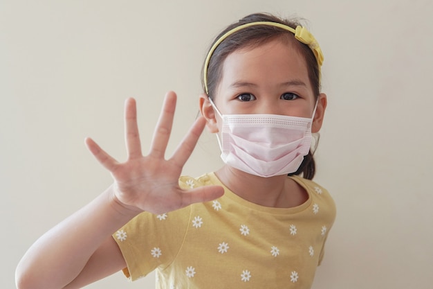 Niña asiática con mascarilla médica y haciendo señal de stop, auto-cuarentena, coronavirus, epidemia de brote de virus covid-19 pandemia.