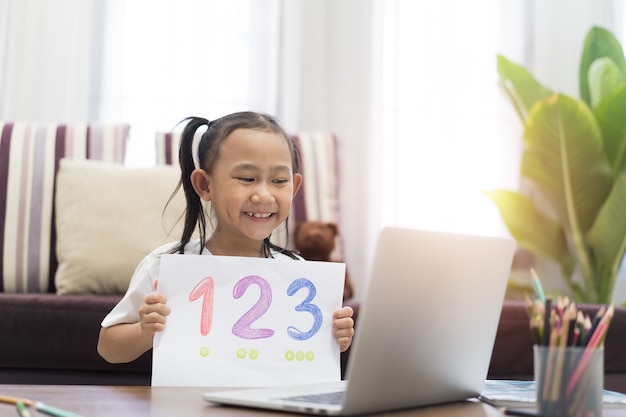 Niña asiática muestra el aprendizaje de la tarea usando la computadora portátil, internet virtual en línea en casa