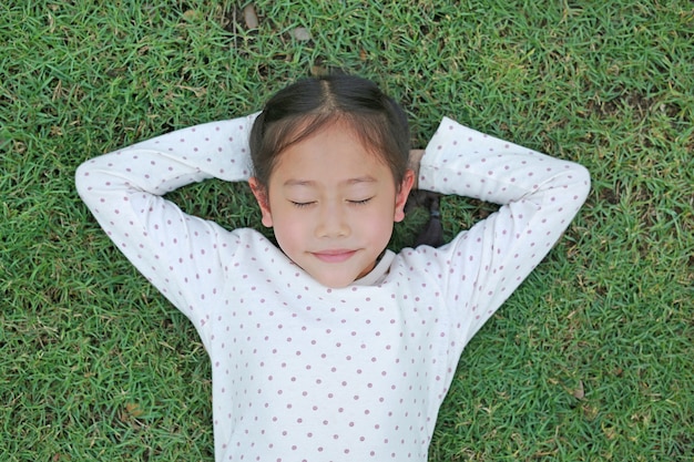 Niña asiática niño sonriendo con las manos detrás de la cabeza y los ojos cerrados descansando sobre la hierba verde al aire libre en el parque de verano. Retrato de vista superior