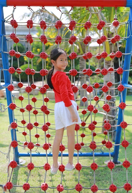 Niña asiática en el patio de recreo jugando en la red de cuerda de escalada Vista posterior