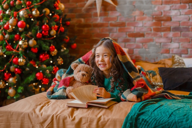 Niña asiática en pijama en el dormitorio acostado en la cama cerca del árbol de Navidad leyendo un libro