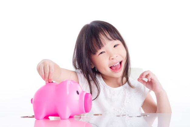 Niña asiática poniendo monedas en hucha rosa sobre fondo blanco