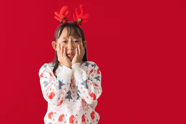 Foto niña asiática positiva con sombrero de papá noel con un suéter navideño mirando sorpresa de pie posando aislada sobre fondo rojo