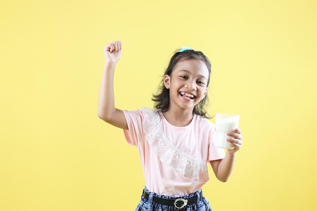 Foto niña asiática sonríe sosteniendo un vaso de leche y muestra poder con un gesto fuerte