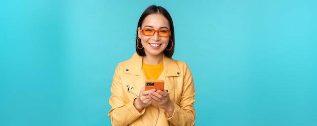 Niña asiática sonriente con gafas de sol usando una aplicación de teléfono inteligente sosteniendo un teléfono móvil de pie sobre fondo azul