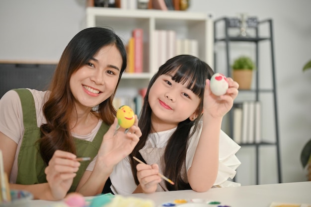 Niña asiática sonriente y su madre mostrando sus huevos de Pascua pintando huevos de Pascua en casa