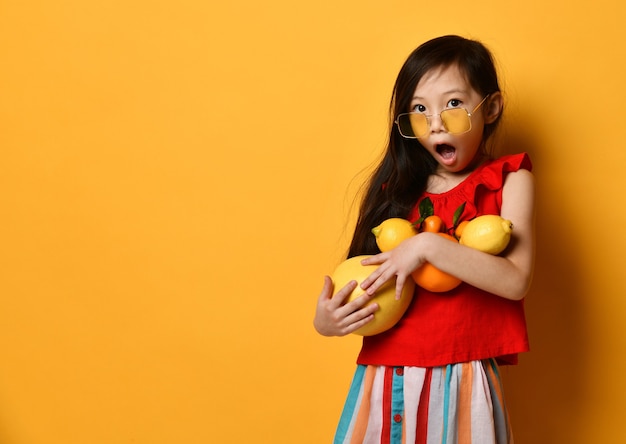 Niña asiática sorprendida en gafas de sol, boina marrón, blusa roja, falda de color. Emocionada, sosteniendo pomelo, naranjas y limones en sus manos, posando sobre fondo naranja. Infancia, frutos.