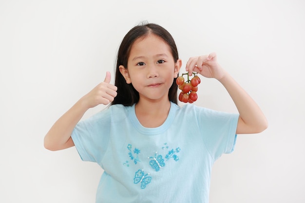 Niña asiática sosteniendo uva morada fresca y haciendo bien firmar pulgar hacia arriba con los dedos sobre fondo blanco.