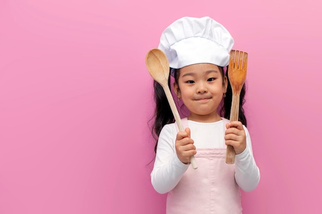 Foto niña asiática en uniforme de chef sosteniendo artículos de cocina sobre fondo rosa aislado