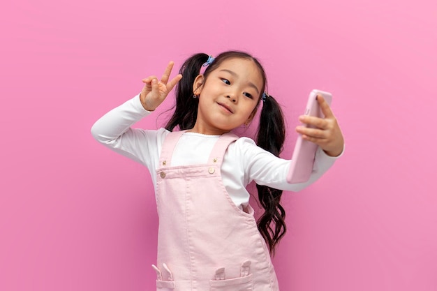 Una niña asiática con un vestido rosa se toma un selfie en un teléfono inteligente y muestra un gesto de paz