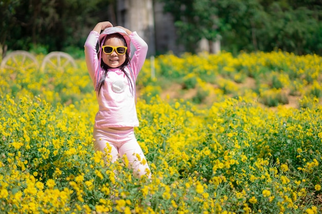 Niña asiática en vestido rosa usar sombrero y gafas de sol de pie en el jardín de flores amarillas