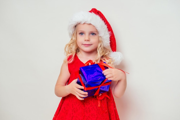 Niña asombrada y sorprendida en un vestido de Navidad gorro de Papá Noel sosteniendo un regalo y globos multicolores de helio. Navidad y año nuevo día de fiesta mágica mañana