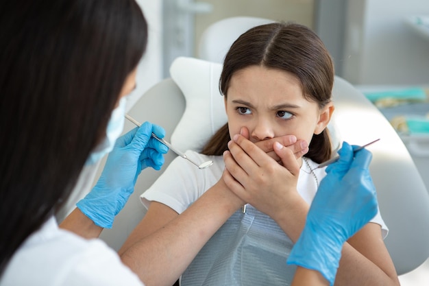 Niña asustada sentada en una silla dental cubriendo la boca con las manos