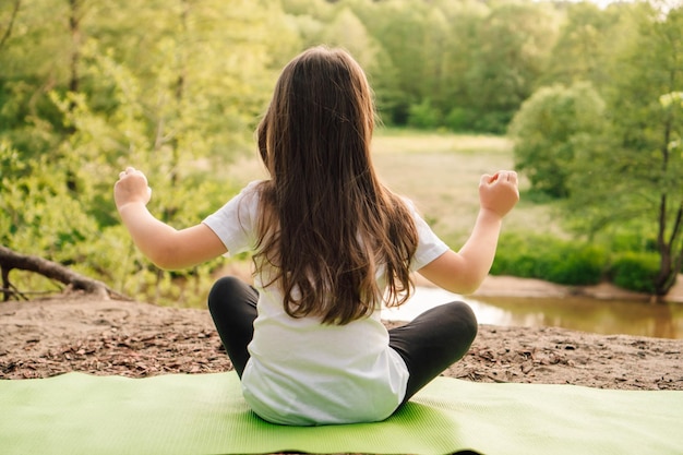 Niña atlética sentada en una alfombra deportiva y practicando yoga frente al río Niño activo meditando en la naturaleza