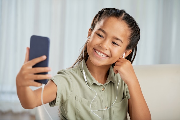 Niña en auriculares sonriendo a la cámara mientras hace selfie en su teléfono móvil