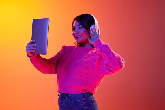 Niña en auriculares con videollamada en línea en tableta sobre fondo naranja degradado en luz de neón Concepto de emociones estilo de vida juvenil