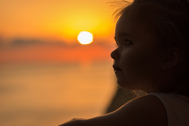 Una niña en el balcón con vistas al mar y al atardecer. Silueta contra el sol.