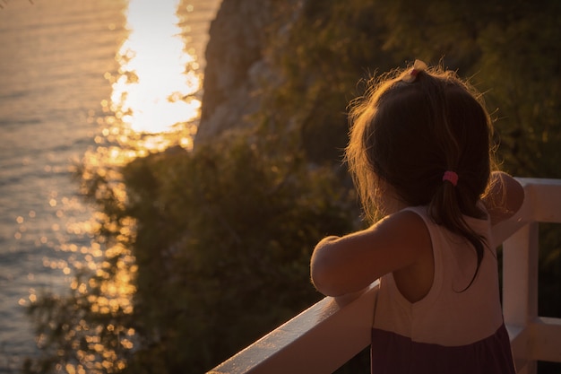 Una niña en el balcón con vistas al mar y al atardecer.