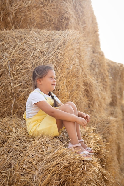 Una niña bastante pensante descansando en un pajar con una leve sonrisa en la cara mirando hacia otro lado y sosteniendo heno en las manos usando un vestido de verano Divirtiéndose lejos de la ciudad en un campo lleno de heno dorado