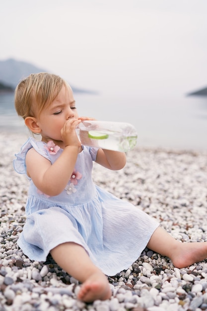 Niña bebe agua de una botella en una playa de guijarros