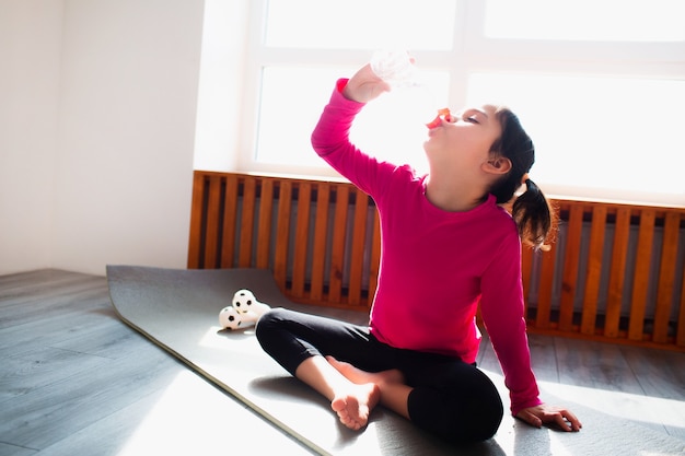 Niña bebe agua después del entrenamiento en casa. Niño lindo está entrenando sobre una alfombra en el interior. La pequeña modelo de mujer de pelo oscuro en ropa deportiva tiene ejercicios cerca de la ventana de su habitación.