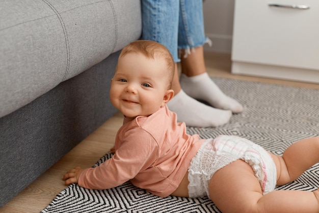 Niña bebé feliz gateando en el piso de la sala de estar en la alfombra cerca del sofá, mirando a la cámara con una sonrisa de niño lindo y encantador, infancia feliz.