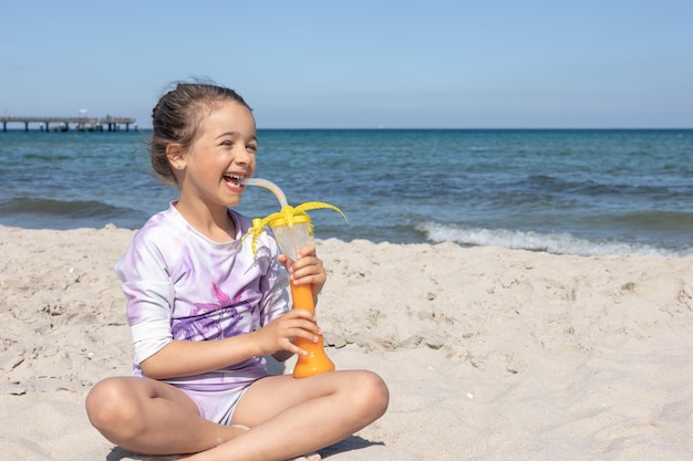 La niña bebe jugo sentada en la arena cerca del mar