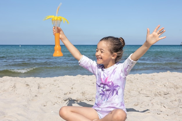 La niña bebe jugo sentada en la arena cerca del mar