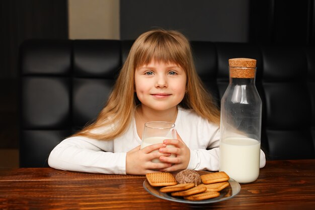 Foto la niña bebe leche con galletas por la mañana en el desayuno. botella con leche y vidrio, galletas en un plato sobre una mesa de madera, desayuno rústico