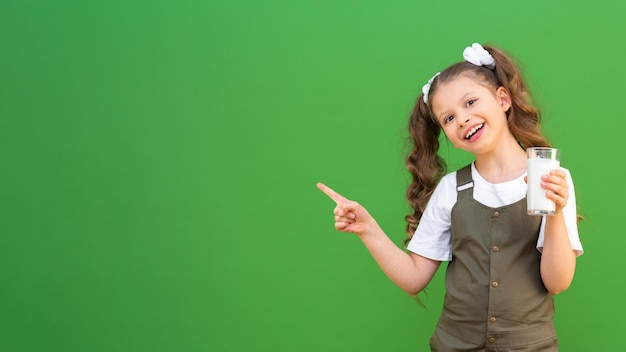 Una niña bebe leche de vaca y señala con el dedo un anuncio sobre un fondo verde aislado. Calcio para el cuerpo del niño, copie el espacio.