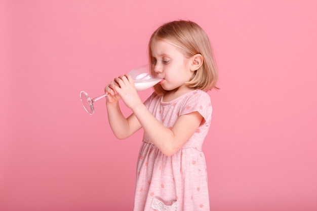 Niña bebe leche de un vaso sobre una superficie rosa