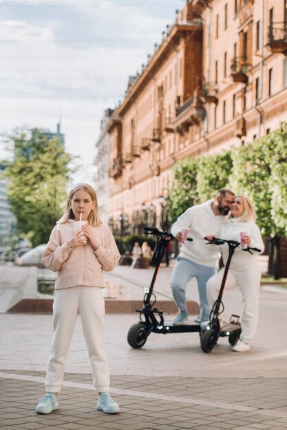 Una niña con una bebida en sus manos está parada en la ciudad y detrás de ella en patinetes eléctricos están sus padres.