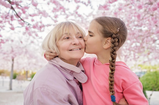 La niña besa a su abuela cerca de la floreciente rama de sakura durante una caminata en el jardín de primavera