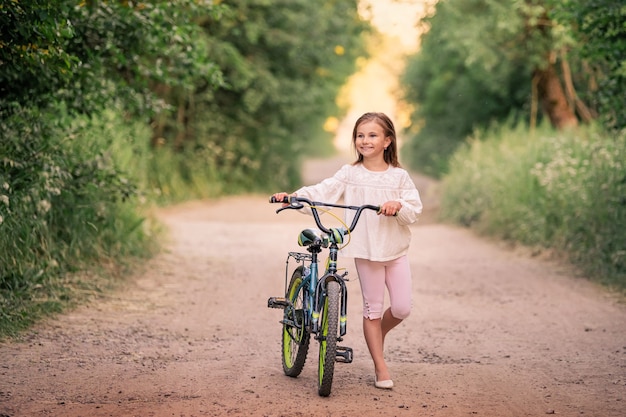 Niña va con una bicicleta por un camino rural en la naturaleza al atardecer