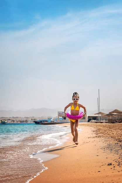 Una niña con un bikini amarillo y un inflable de anillo rosa corre a lo largo de la playa.