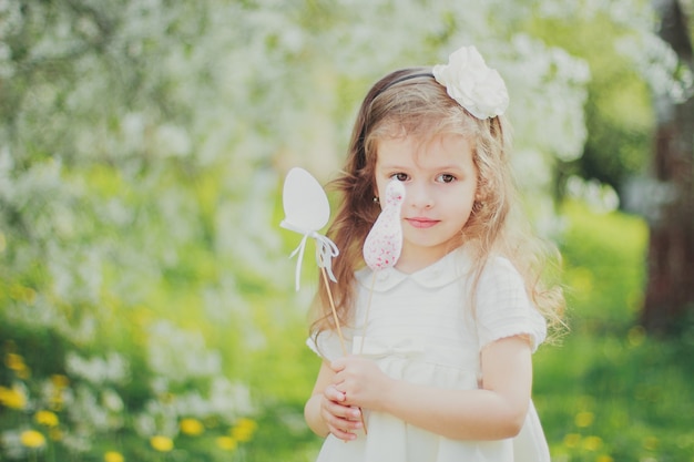 Niña en blanco mantenga huevos de pascua de juguete en palos en el jardín de cerezos de primavera