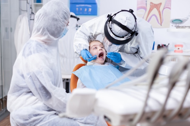 Niña con la boca abierta recibiendo tratamiento de caries de dentista pediátrico vestido con traje de ppe. Dentista en traje de coronavirus con espejo curvo durante el examen de los dientes del niño.