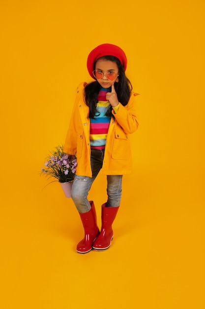 una niña con una boina roja con gafas rosas en un impermeable amarillo sosteniendo un cubo con flores