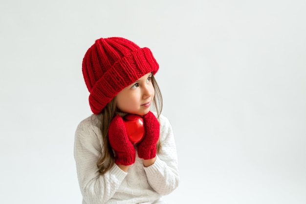 Una niña con una bola de Navidad en un gorro de punto rojo y guantes mirar a un lado en el espacio de la copia