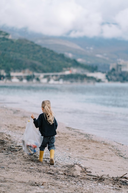 Niña con una bolsa de basura camina por la playa a lo largo de la vista trasera del mar