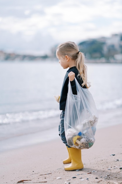 Niña con una bolsa de basura de pie en la playa y mira al mar