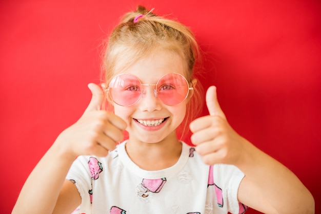 Niña bonita alegre en gafas rosas mostrando los pulgares hacia arriba sobre rojo