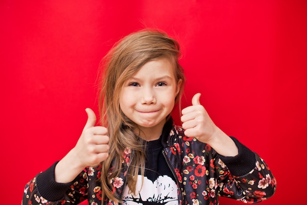 Niña bonita alegre que muestra los pulgares para arriba sobre la pared roja