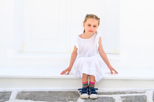 Niña bonita y blong con un vestido blanco sentada al aire libre cerca de la casa blanca