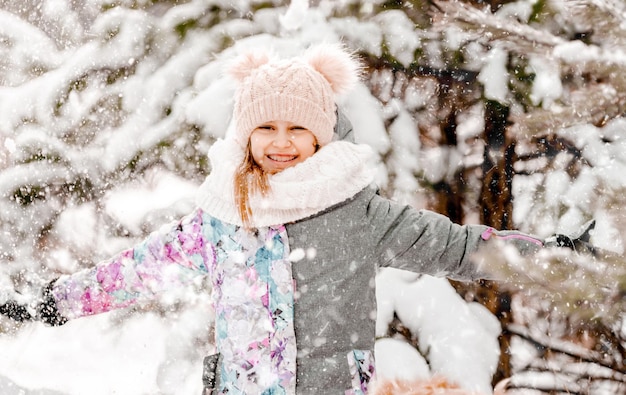 Niña bonita en el bosque nevado en invierno mirando a la cámara y sonriendo con copos de nieve en el aire. Niña preadolescente con sombrero en retrato de clima frío