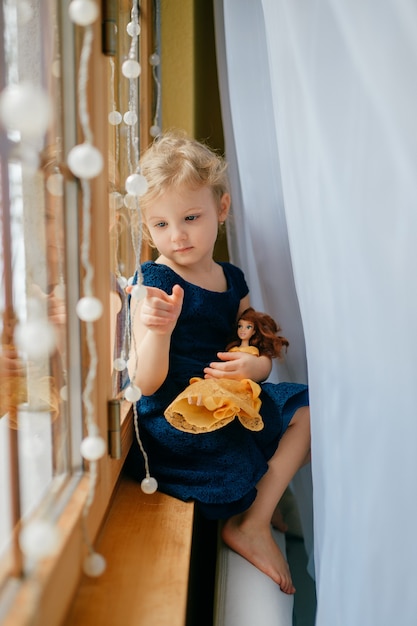 Niña bonita con cabello rubio vestido azul sostiene su precioso juguete y se sienta en la habitación del bebé en un alféizar y sonríe