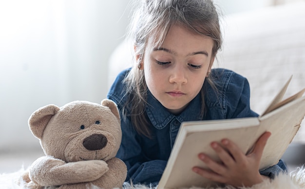 Niña bonita en casa, tirada en el suelo con su juguete favorito y lee el libro.